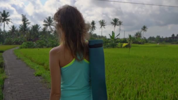 Slowmotion Steadicam shot van een jonge vrouw met yoga math wandelen door een prachtige rijstveld — Stockvideo