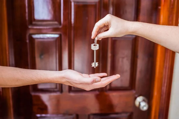 Mano femenina dando una llave a los hombres de la casa en el fondo de la puerta de madera. Poseer concepto de bienes raíces — Foto de Stock