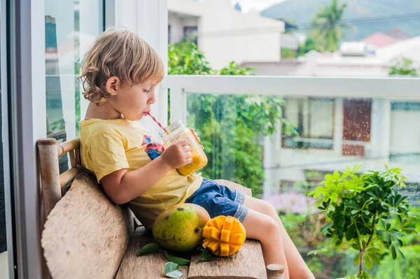 Chico bebiendo jugoso batido de mango en tarro de cristal de albañil con paja roja rayada sobre fondo de madera vieja. Concepto de vida saludable, espacio de copia — Foto de Stock