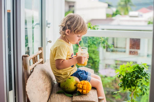 Chico bebiendo jugoso batido de mango en tarro de cristal de albañil con paja roja rayada sobre fondo de madera vieja. Concepto de vida saludable, espacio de copia — Foto de Stock