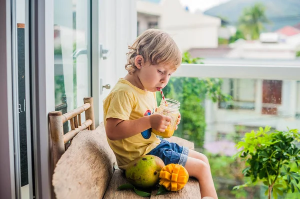 Chico bebiendo jugoso batido de mango en tarro de cristal de albañil con paja roja rayada sobre fondo de madera vieja. Concepto de vida saludable, espacio de copia — Foto de Stock