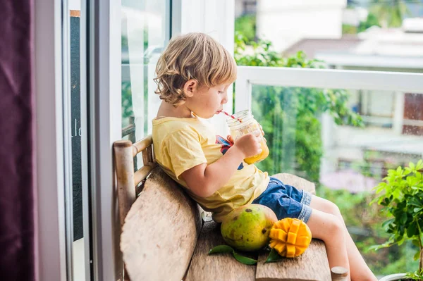 Chico bebiendo jugoso batido de mango en tarro de cristal de albañil con paja roja rayada sobre fondo de madera vieja. Concepto de vida saludable, espacio de copia — Foto de Stock