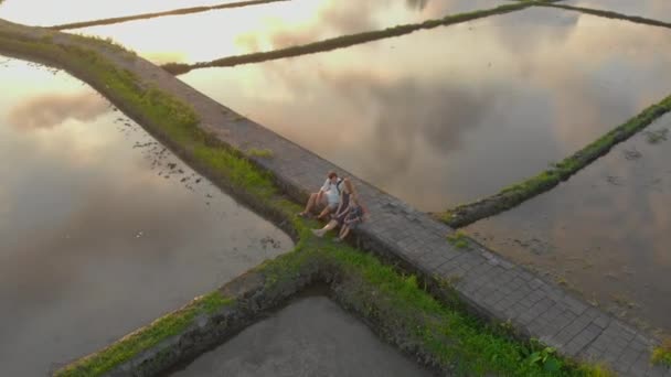 Aerial shot of a happy family that is sitting on a walkway through the big field filled by water. They enjoy sunset time and clouds reflecting in water. Travel to Bali concept — Stock Video