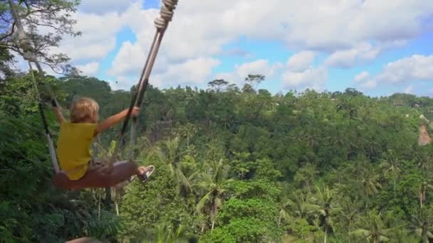 El niño se balancea en columpios en un entorno tropical. Viajes al concepto de Bali — Vídeos de Stock