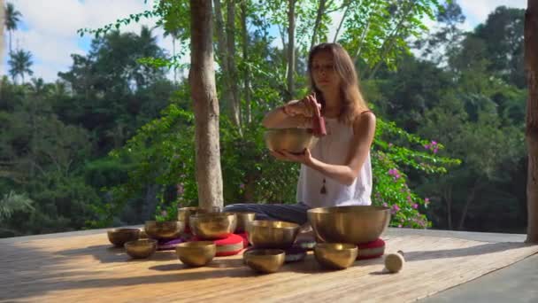 Woman playing on Tibetan singing bowl while sitting on yoga mat against a waterfall — Stock Video