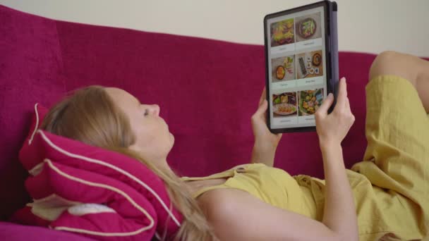 La jeune femme commande de la nourriture pour le déjeuner en ligne à l'aide d'une tablette. Elle est allongée sur un canapé dans son appartement — Video