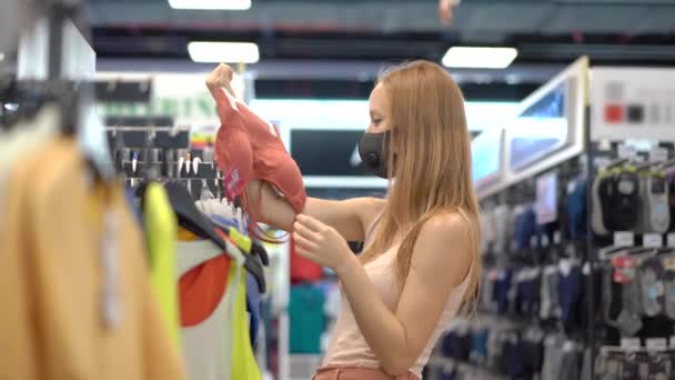 Mujer en una tienda de ropa con una máscara médica por un coronovirus. La cuarentena ha terminado, ahora puedes ir a la tienda de ropa pero tienes que usar una mascarilla. — Vídeo de stock