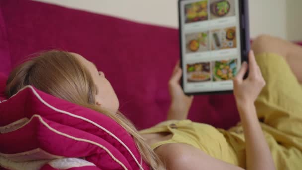 Giovane donna ordina cibo per il pranzo online utilizzando un tablet. Lei è sdraiata su un divano nel suo appartamento — Video Stock