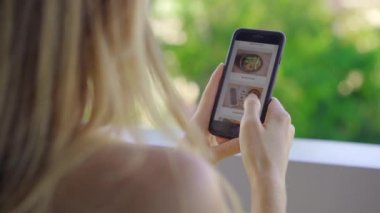 Genç bir kadın akıllı telefon kullanarak öğle yemeği siparişi veriyor. Dairesinde bir balkonda oturuyor.