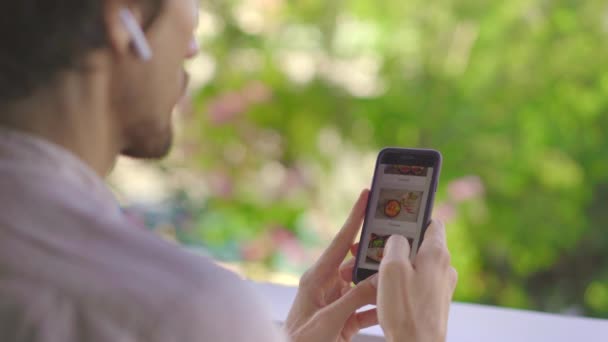 Jovem pede comida para o almoço online usando um smartphone. Ele está sentado em uma varanda em seu apartamento — Vídeo de Stock