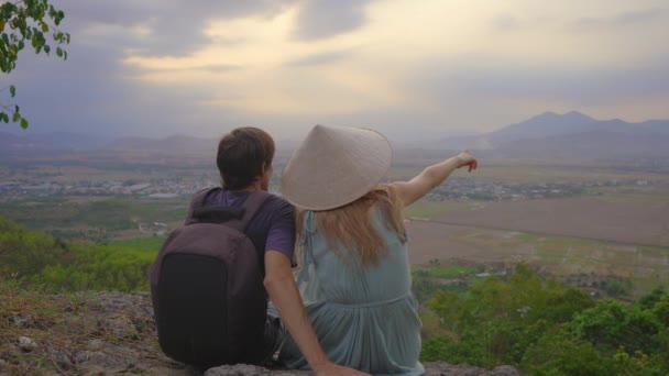 Una joven pareja de turistas se sientan en la cima de una montaña con una vista increíble de un valle. Ella lleva un sombrero asiático — Vídeos de Stock
