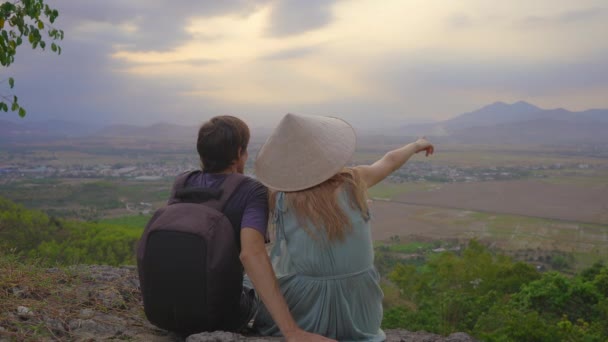 Mladý pár turistů sedí na vrcholku hory s úžasným výhledem na údolí. Nosí asijský klobouk. — Stock video