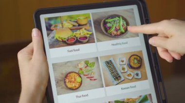Genç bir kadın, tablet kullanarak internetten yemek sipariş ediyor. Dairesinde bir mutfakta oturuyor.