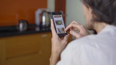 Genç adam akıllı telefon kullanarak internetten yemek sipariş ediyor. Dairesinde bir mutfakta oturuyor.