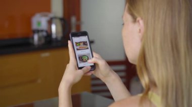 Genç bir kadın akıllı telefon kullanarak internetten yemek sipariş ediyor. Dairesinde, mutfağında oturuyor.