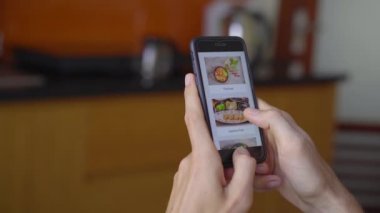 Genç adam akıllı telefon kullanarak internetten yemek sipariş ediyor. Dairesinde bir mutfakta oturuyor.