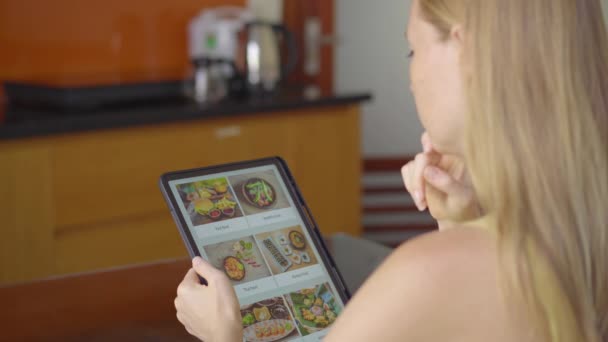 Giovane donna ordina cibo per il pranzo online utilizzando un tablet. Lei è seduta in una cucina nel suo appartamento — Video Stock