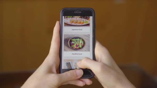 Une jeune femme commande de la nourriture en ligne à l'aide d'un smartphone. Elle est assise dans sa cuisine dans son appartement — Video