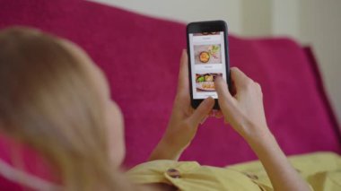 Genç bir kadın akıllı telefon kullanarak internetten yemek sipariş ediyor. Dairesinde bir kanepede yatıyor.