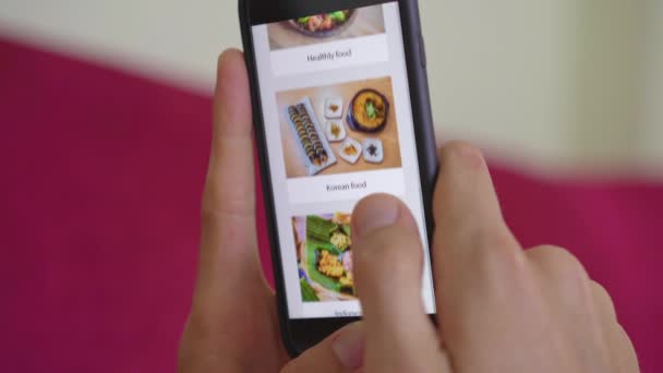 Giovane uomo ordina cibo per il pranzo online utilizzando uno smartphone. Lui è sdraiato su un divano nel suo appartamento — Video Stock