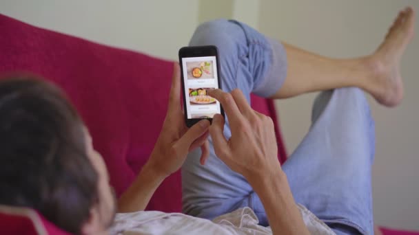 Unge man beställer mat till lunch på nätet med hjälp av en smartphone. Han ligger på en soffa i sin lägenhet. — Stockvideo