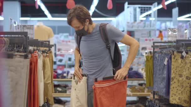 Un hombre en una tienda de ropa con una máscara médica por un coronovirus. La cuarentena ha terminado, ahora puedes ir a la tienda de ropa pero tienes que usar una mascarilla. — Vídeo de stock