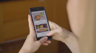Genç bir kadın akıllı telefon kullanarak internetten yemek sipariş ediyor. Dairesinde, mutfağında oturuyor.