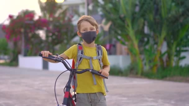 Actieve schooljongen met medisch masker op een fiets met rugzak op zonnige dag. Gelukkig kind fietsen op weg naar school. Je moet met een masker naar school vanwege de coronavirus epidemie. — Stockvideo