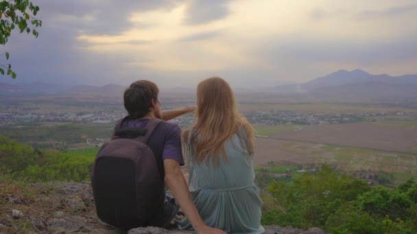 Um jovem casal de turistas se senta em um topo de montanha com uma vista incrível sobre um vale — Vídeo de Stock