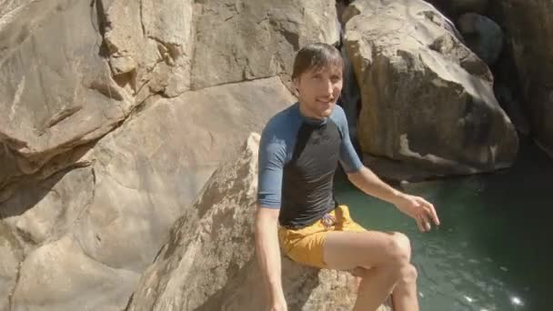 Plan au ralenti d'un jeune homme qui s'enregistre assis sur une falaise au bord d'une rivière tropicale avec une cascade. Concept d'été — Video