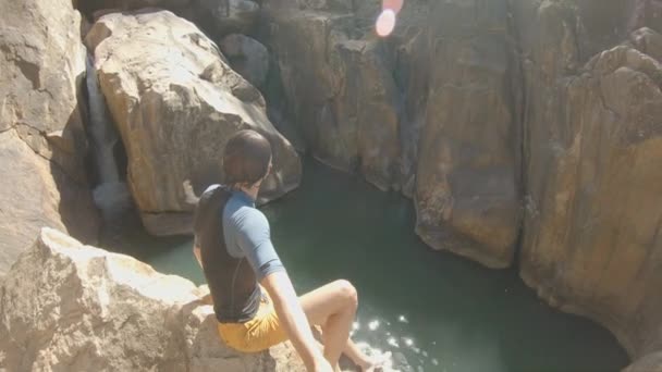 Plan au ralenti d'un jeune homme qui s'enregistre assis sur une falaise au bord d'une rivière tropicale avec une cascade. Concept d'été — Video