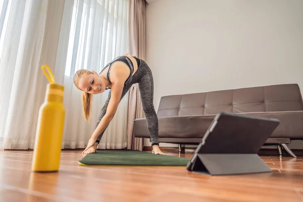 健身女人在家里的地板上锻炼，并在平板电脑上观看健身录像。人们在网上做运动是因为这种病毒 — 图库照片