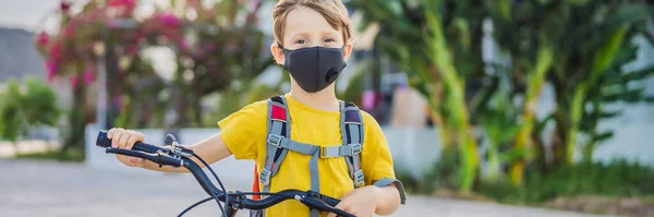 阳光明媚的日子里，戴着医疗面罩的积极的学童骑着背包骑自行车。快乐的孩子骑自行车上学的路上。你需要带着面具去学校，因为在朗，卡洛纳韦病毒正在流行 — 图库照片