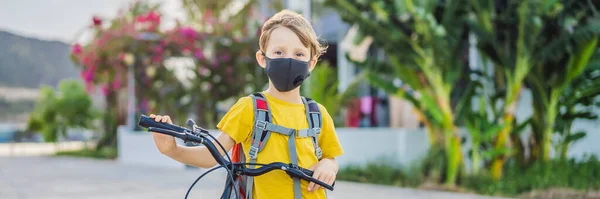 Aktív iskolás fiú orvosi maszkban biciklizik hátizsákkal a napsütéses napon. Boldog gyerek biciklizés úton az iskolába. Maszkban kell iskolába menned a koronavírus járvány miatt, BANNER, LONG — Stock Fotó