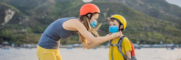 Активный школьник и его мама в медицинской маске и защитном шлеме едут на велосипеде с рюкзаком в солнечный день. Счастливый ребенок едет в школу на велосипеде. Вы должны пойти в школу в маске из-за — стоковое фото