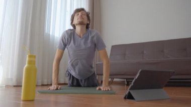 Evde yoga yapan genç bir adam tabletten izlediği bir videonun talimatlarını takip ediyor. Sosyal uzaklık kavramı. İnternet çalıştırıcı kavramı