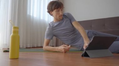 Evde yoga yapan genç bir adam tabletten izlediği bir videonun talimatlarını takip ediyor. Sosyal uzaklık kavramı. İnternet çalıştırıcı kavramı