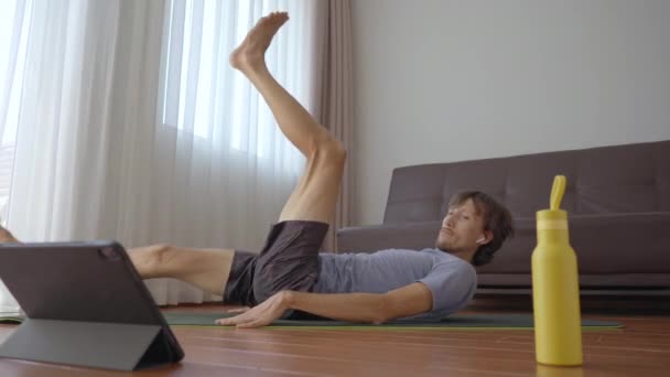 Ein junger Mann zu Hause macht körperliche Übungen nach Anweisungen aus einem Video, das er sich auf einem Tablet ansieht. Soziale Distanzierung. Internet-Trainer-Konzept — Stockvideo
