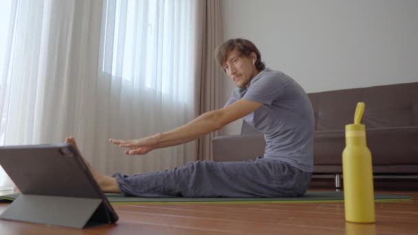 Młody człowiek w domu ćwiczy jogę, wykonując instrukcje z filmu, który ogląda na tablecie. Koncepcja dystansu społecznego. Koncepcja trenera internetowego — Wideo stockowe