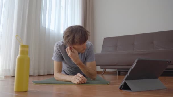 Un joven en casa haciendo yoga siguiendo las instrucciones de un vídeo que ve en una tableta. Concepto de distanciamiento social. Concepto de entrenador de Internet — Vídeos de Stock