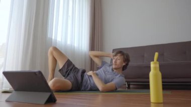 Tabletten izlediği bir videoda talimatları takip eden genç bir adam evde fiziksel egzersiz yapıyor. Sosyal uzaklık kavramı. İnternet çalıştırıcı kavramı