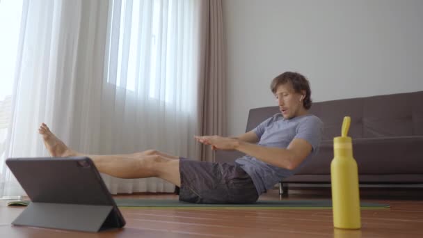 Un jeune homme à la maison faisant des exercices physiques suivant les instructions d'une vidéo qu'il regarde sur une tablette. Concept de distance sociale. Concept de formateur Internet — Video