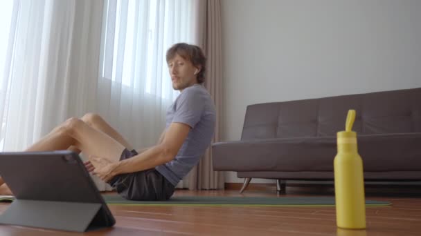 Młody mężczyzna w domu ćwiczy fizycznie zgodnie z instrukcjami z filmu, który ogląda na tablecie. Koncepcja dystansu społecznego. Koncepcja trenera internetowego — Wideo stockowe