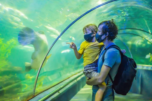 Pa en zoon met medische maskers kijken naar de vissen in het aquarium in het oceanarium. Mensen lopen in medische maskers na het einde van het coronovirus — Stockfoto