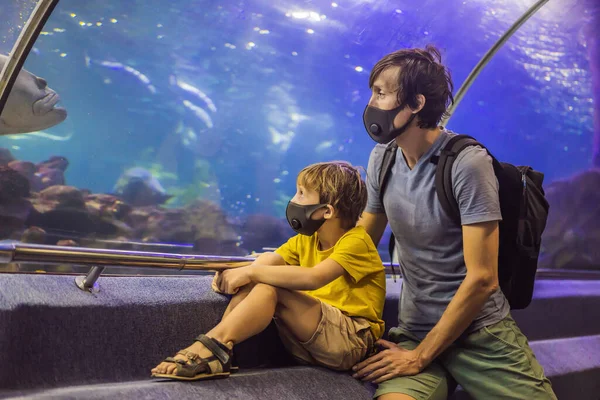 Ο μπαμπάς και ο γιος με ιατρικές μάσκες κοιτούν τα ψάρια στο ενυδρείο στο ωκεάνιο. Οι άνθρωποι περπατούν με ιατρικές μάσκες μετά το τέλος του κορονοϊού. — Φωτογραφία Αρχείου