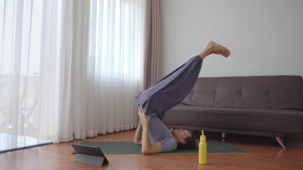 Ein junger Mann macht zu Hause Yoga nach Anweisungen aus einem Video, das er sich auf einem Tablet ansieht. Soziale Distanzierung. Internet-Trainer-Konzept — Stockvideo