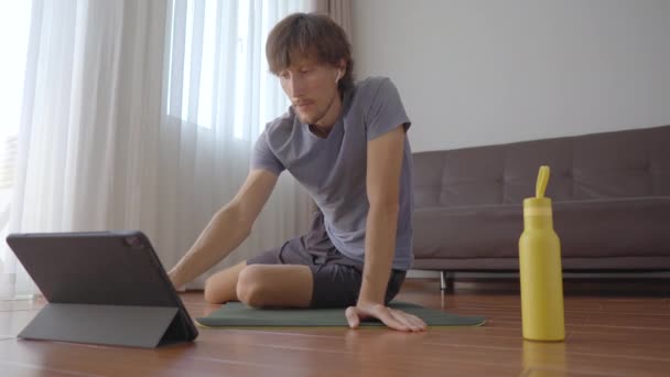 Młody mężczyzna w domu ćwiczy fizycznie zgodnie z instrukcjami z filmu, który ogląda na tablecie. Koncepcja dystansu społecznego. Koncepcja trenera internetowego — Wideo stockowe