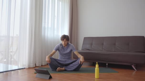 家でヨガをしている若い男が、タブレットで見ているビデオの指示に従ってヨガをしている。社会的距離概念。インターネットトレーナーの概念 — ストック動画