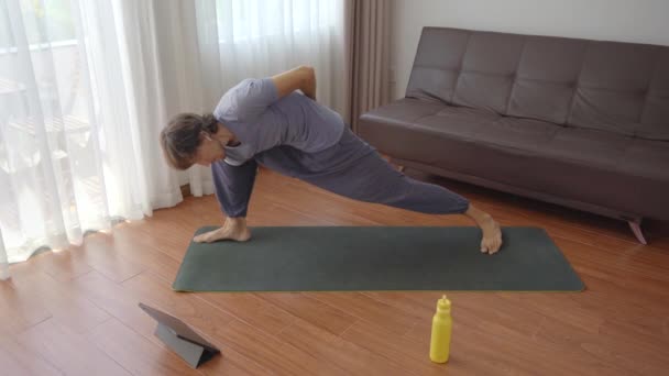 Um jovem em casa fazendo ioga seguindo as instruções de um vídeo que ele assiste em um tablet. Conceito de distanciamento social. Conceito de formador de Internet — Vídeo de Stock