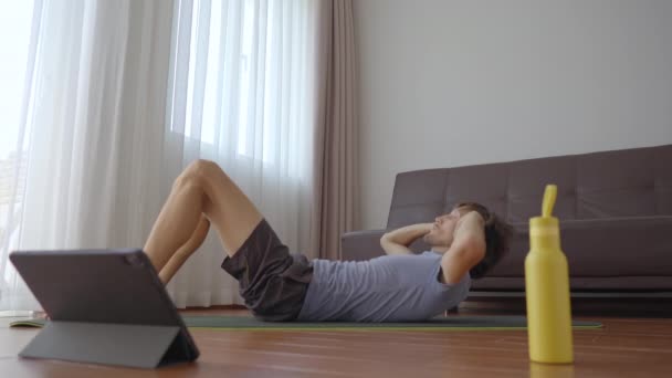 Een jongeman thuis doet fysieke oefeningen volgens instructies van een video die hij bekijkt op een tablet. Sociaal afstandelijk concept. Het concept van internettrainer — Stockvideo